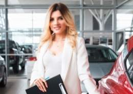 Friendly car salesperson standing next to new vehicle in dealership showroom.