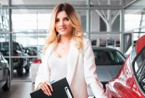 Friendly car salesperson standing next to new vehicle in dealership showroom.