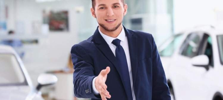 Friendly car salesman handshake at dealership, what you say next at the dealership could cost you. Car salesman greeting customer at dealership – car shopping negotiation begins.