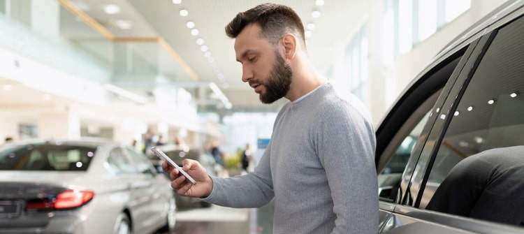 Car buyer in denial comparing car prices during dealership visit. Man at car dealership in car buyer denial checking prices on phone.